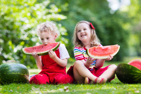 Child eating watermelon in the garden. Kids eat fruit outdoors. Healthy snack for children. Little girl and boy playing in the garden biting a slice of water melon.の写真素材