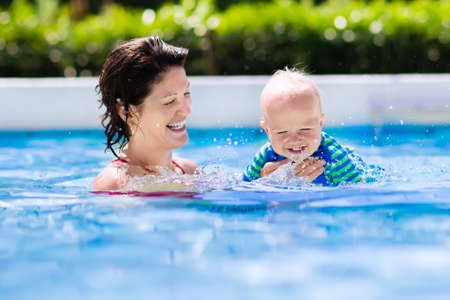 Happy young mother playing with her baby in outdoor swimming pool on hot summer day. Kids learn to swim during family vacation. Children wearing sun protection rash guard relaxing in tropical resortの写真素材