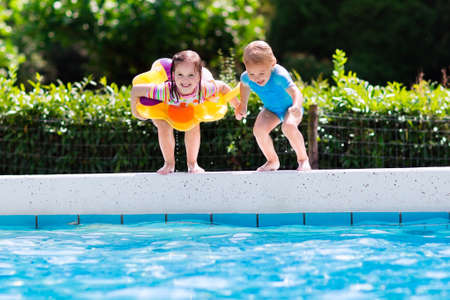 Happy little girl and boy holding hands jumping into outdoor swimming pool in a tropical resort during family summer vacation. Kids learning to swim. Water fun for children.の写真素材