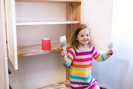 Happy preschool age girl holding a brush painting a wooden bedroom closet with white color paint. Child learning furniture renovation and restoration. Do it yourself project with kids at home.の写真素材