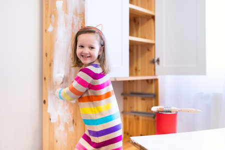 Happy preschool age girl holding a brush painting a wooden bedroom closet with white color paint. Child learning furniture renovation and restoration. Do it yourself project with kids at home.の写真素材