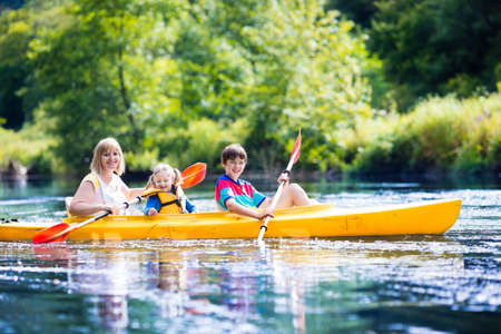 Happy family with two kids enjoying kayak ride on beautiful river. Mother with little girl and teenager boy kayaking on hot summer day. Water sport fun. Canoe and boat for children.の写真素材