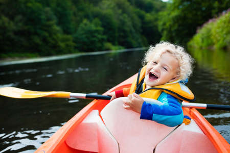 Happy kid enjoying kayak ride on beautiful river. Little curly toddler boy kayaking on hot summer day. Water sport and camping fun. Canoe for children. Funny child with vessel in a boat.の写真素材
