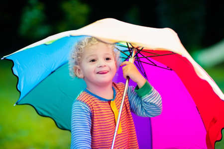 Little boy playing in rainy summer park. Child with colorful rainbow umbrella standing in the rain. Kid walking in autumn shower. Outdoor fun by any weatherの写真素材