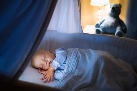 Adorable baby sleeping in blue bassinet with canopy at night. Little boy in pajamas taking a nap in dark room with crib, lamp and toy bear. Bed time for kids. Bedroom and nursery interior.の写真素材