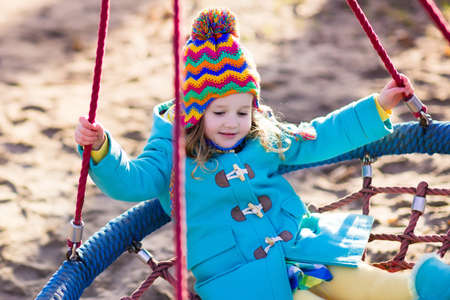 Little girl in warm coat and hat on a playground. Child playing outdoors in autumn. Kids play on school yard. Happy kid in kindergarten or preschool. Children having fun on cold fall day. Toddler on a swing.の写真素材