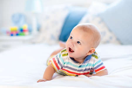 Adorable baby boy in white sunny bedroom. Newborn child relaxing in bed. Nursery for young children....