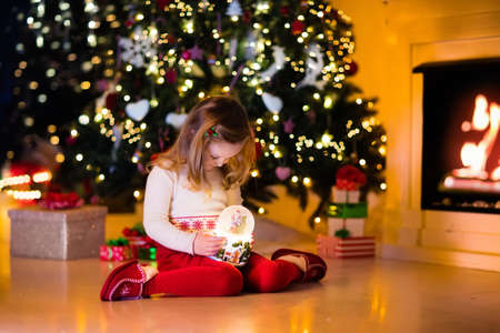 Family on Christmas eve at fireplace. Little girl opening Xmas presents holding snow globe. Child under Christmas tree with gift boxes. Decorated living room. Cozy winter evening at home.の写真素材