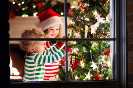 Happy little kids in matching red and green striped pajamas decorate Christmas tree in beautiful living room with traditional fire place. Children opening presents on Xmas eve. View though window.の写真素材