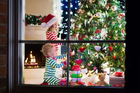 Happy little kids in matching red and green striped pajamas decorate Christmas tree in beautiful living room with traditional fire place. Children opening presents on Xmas eve.の写真素材