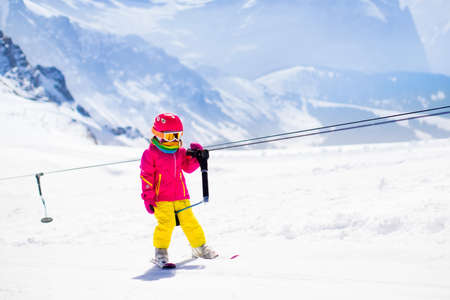 Child on a button ski lift going uphill in the mountains on a sunny snowy day. Kids in winter sport school in alpine resort. Family fun in the snow. Little skier learning and exercising on a slope.の写真素材