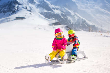 Little girl and baby boy play with snow on a sleigh ride in Swiss Alps mountains. Children sledding on sunny and snowy winter day. Toddler kids riding a sledge in mountain landscape of Switzerland.の写真素材