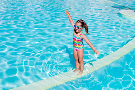Happy laughing little girl playing in outdoor swimming pool on a hot summer day. Kid in colorful bathing suit and goggles learning to swim in tropical resort. Water fun for children.の写真素材