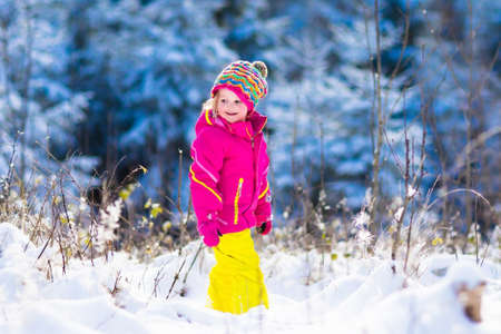Child running in snowy forest. Toddler kid playing outdoors. Kids play in snow. Christmas vacation in sunny winter park for family with young children. Little girl in colorful jacket and knitted hat.の写真素材