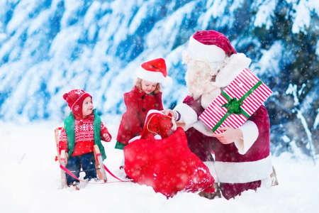 Santa Claus and children opening presents in snowy forest. Kids and father in Santa costume and beard open Christmas gifts. Little girl helping with present sack. Xmas, snow and winter fun for family.の写真素材
