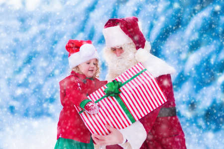 Santa Claus and children opening presents in snowy forest. Kids and father in Santa costume and beard open Christmas gifts. Little girl helping with present sack. Xmas, snow and winter fun for family.の写真素材