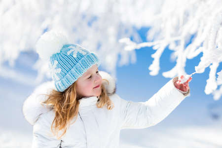 Child in white jacket with fur hood and blue knitted snowflake hat playing in snowy forest on sunny winter day. Toddler kid girl having fun outdoors during Christmas vacation. Kids play in snow.の写真素材