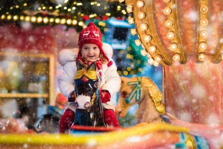 Happy little girl in warm jacket and red knitted Nordic hat and scarf riding carousel horse during family trip to traditional German Christmas market. Kids at Xmas outdoor fair on snowy winter day.の写真素材