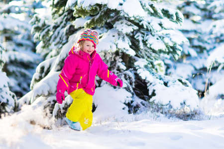 Child running in snowy forest. Toddler kid playing outdoors. Kids play in snow. Christmas vacation in sunny winter park for family with young children. Little girl in colorful jacket and knitted hat.の写真素材