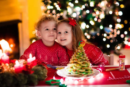 Little boy and girl making Christmas gingerbread house at fireplace in decorated living room. Kids playing with ginger bread under Christmas tree. Baking and cooking with children for Xmas at home.の写真素材
