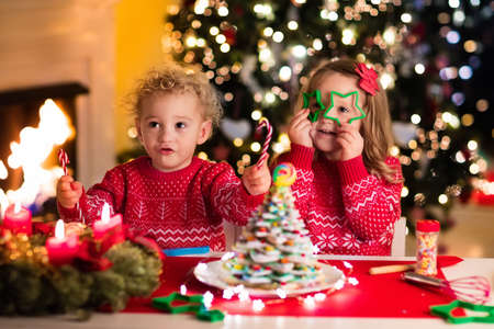 Little boy and girl making Christmas gingerbread house at fireplace in decorated living room. Kids playing with ginger bread under Christmas tree. Baking and cooking with children for Xmas at home.の写真素材