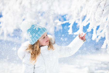 Child in white jacket with fur hood and blue knitted snowflake hat playing in snowy forest on sunny winter day. Toddler kid girl having fun outdoors during Christmas vacation. Kids play in snow.の写真素材
