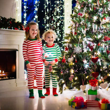 Happy little kids in matching red and green striped pajamas decorate Christmas tree in beautiful living room with traditional fire place. Children opening presents on Xmas eve.の写真素材