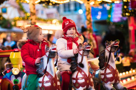 Happy little girl and boy in warm jacket and red knitted Nordic hat and scarf riding carousel horse during family trip to traditional German Christmas market. Kids at Xmas outdoor fair on snowy winter day.の写真素材