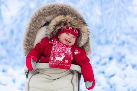 Happy laughing baby in warm red down jacket and knitted Nordic hat and scarf on a walk in a snowy winter park sitting in warm stroller with sheepskin hood catching snow. Child in buggy with foot muffの写真素材