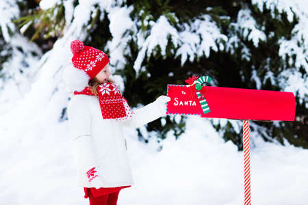 Happy child in knitted reindeer hat and scarf holding letter to Santa with Christmas presents wish list at red mail box in snow under Xmas tree in winter forest. Kids sending post to North Pole.の写真素材