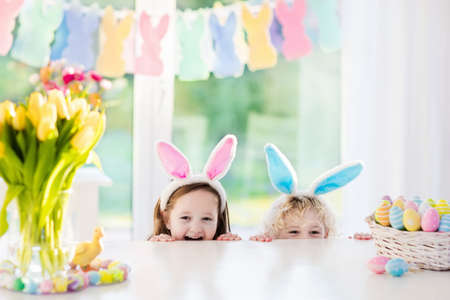 Boy and girl in bunny ears at breakfast on Easter morning at table with Easter eggs basket. Kids celebrating Easter. Children on Easter egg hunt. Home decoration - pastel bunny banner and flowers.の写真素材