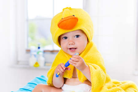 Little baby in yellow duck towel brushing teeth on changing table after bath. Infant boy with tooth brush. Dental hygiene, toothbrush and toothpaste for young kids. Child teeth and oral health care.の写真素材