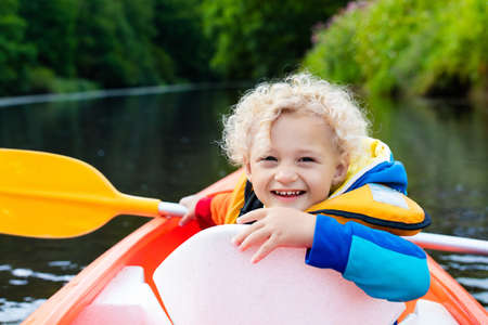 Happy kid enjoying kayak ride on beautiful river. Little curly toddler boy kayaking on hot summer day. Water sport and camping fun. Canoe for children. Funny child with vessel in a boat.の写真素材