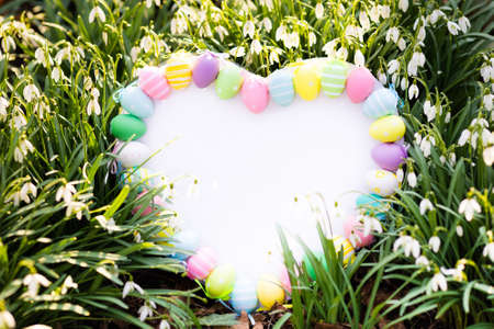 beautiful white board in heart shape decorated with pastel color easter eggs in spring garden with snowdrop flowers copy space for text colorful decoration for easter egg hunt and celebrationの写真素材