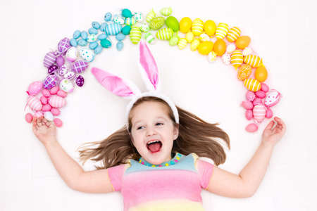 Funny little girl with bunny ears having fun on Easter egg hunt. Child playing with colorful Easter eggs. Kids play with pastel color rainbow eggs. Spring and Easter art and crafts for children.の写真素材