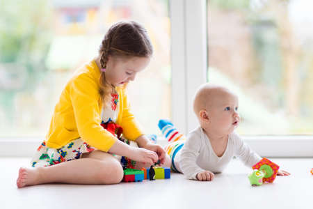 Kids playing together at home. Little girl and boy play sitting on the floor next to a window. Toddler child building tower with colorful blocks. Baby starting to crawl. Toys for young children.の写真素材