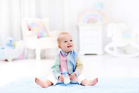 Adorable baby boy playing on a blue floor mat and drinking milk from a bottle in a white sunny...