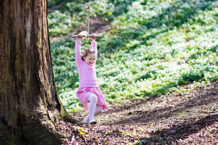 Cute little girl in pink dress on tree rope swing in blooming spring garden with flowers. Child swinging outdoors in backyard playground. Kids play outdoor in summer. Children playing in sunny park.の写真素材