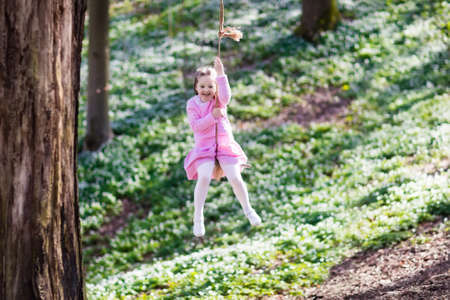 Cute little girl in pink dress on tree rope swing in blooming spring garden with flowers. Child swinging outdoors in backyard playground. Kids play outdoor in summer. Children playing in sunny park.の写真素材