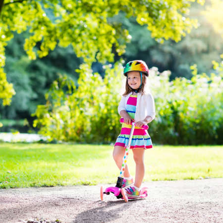 Little child learning to ride a scooter in a city park on sunny summer day. Cute preschooler girl in safety helmet riding a roller. Kids play outdoors. Active leisure and outdoor sport for children.の写真素材