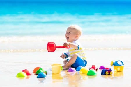 Cute laughing baby boy wearing sun protection rash guard playing with bucket and shovel on tropical beach during family summer sea vacation. Swimwear and beach toys for kids. Child digging in sand.の写真素材