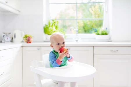 Baby eating fruit. Little boy biting apple sitting in white high chair in sunny kitchen with window and sink. Healthy nutrition for kids. Solid food for infant. Snack or breakfast for young child.の写真素材