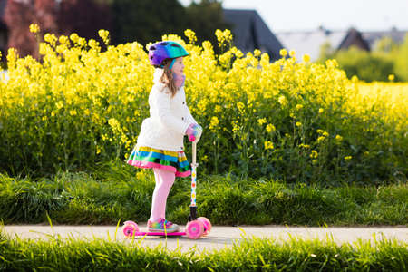 Child riding scooter on way back to school. Little girl playing outdoors learning to balance on kick board. Kids ride scooters in suburbs street. Preschooler in safe helmet on bike or roller.の写真素材