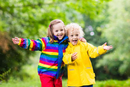 Little boy and girl play in rainy summer park. Children with colorful rainbow jacket and waterproof boots jump in puddle and mud in the rain. Kids walk in autumn shower. Outdoor fun by any weather.の写真素材