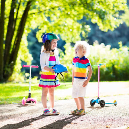 Children learn to ride scooter in a park on summer day. Preschooler girl helping boy to put on safety helmet. Siblings riding roller. Kids play outdoors with scooters. Active outdoor sport for child.の写真素材