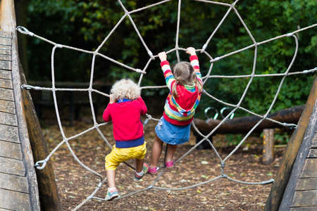 Active children playing on climbing net at school yard playground. Kids play and climb outdoors on sunny summer day. Cute girl and boy on nest swing at preschool sport center. Family summer vacation.の写真素材