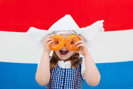 Little Dutch girl wearing traditional national costume, dress and hat holding orange donuts at flag of the Netherlands. Child with Oranje souvenirs from Holland celebrating King day. Kid with sweets.の写真素材