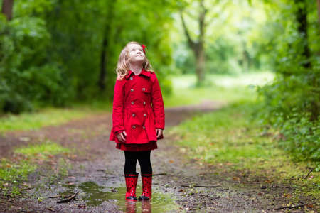 Little girl playing in rainy summer park. Child with red ladybug umbrella, waterproof coat and boots jumping in puddle and mud in the rain. Kid walking in autumn shower. Outdoor fun by any weather.の写真素材