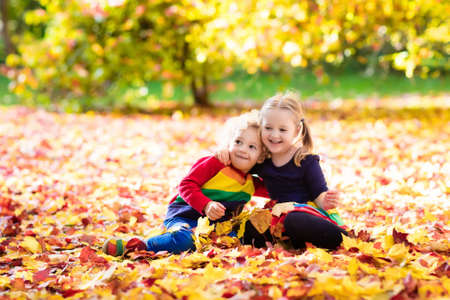 Kids play in autumn park. Children throwing yellow maple leaves. Boy and girl jump and run with oak leaf. Fall foliage. Family outdoor fun in autumn. Toddler kid and preschooler child in fall.の写真素材