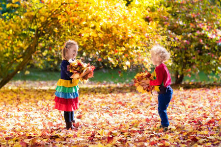 Kids play in autumn park. Children throwing yellow maple leaves. Boy and girl jump and run with oak leaf. Fall foliage. Family outdoor fun in autumn. Toddler kid and preschooler child in fall.の写真素材